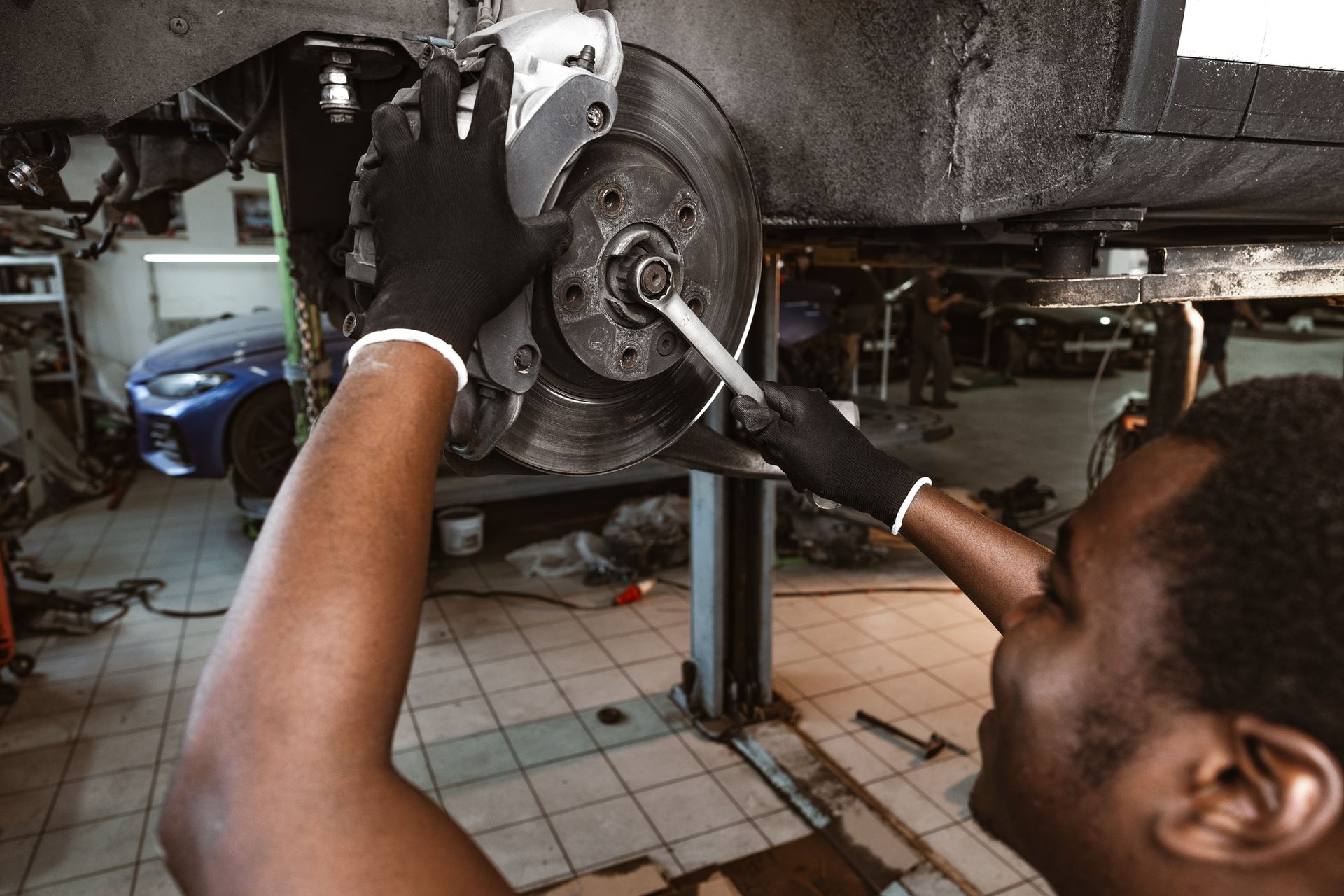 Mechanic working on a car's brakes. Black gloves, wrench, blue car in background, garage setting.