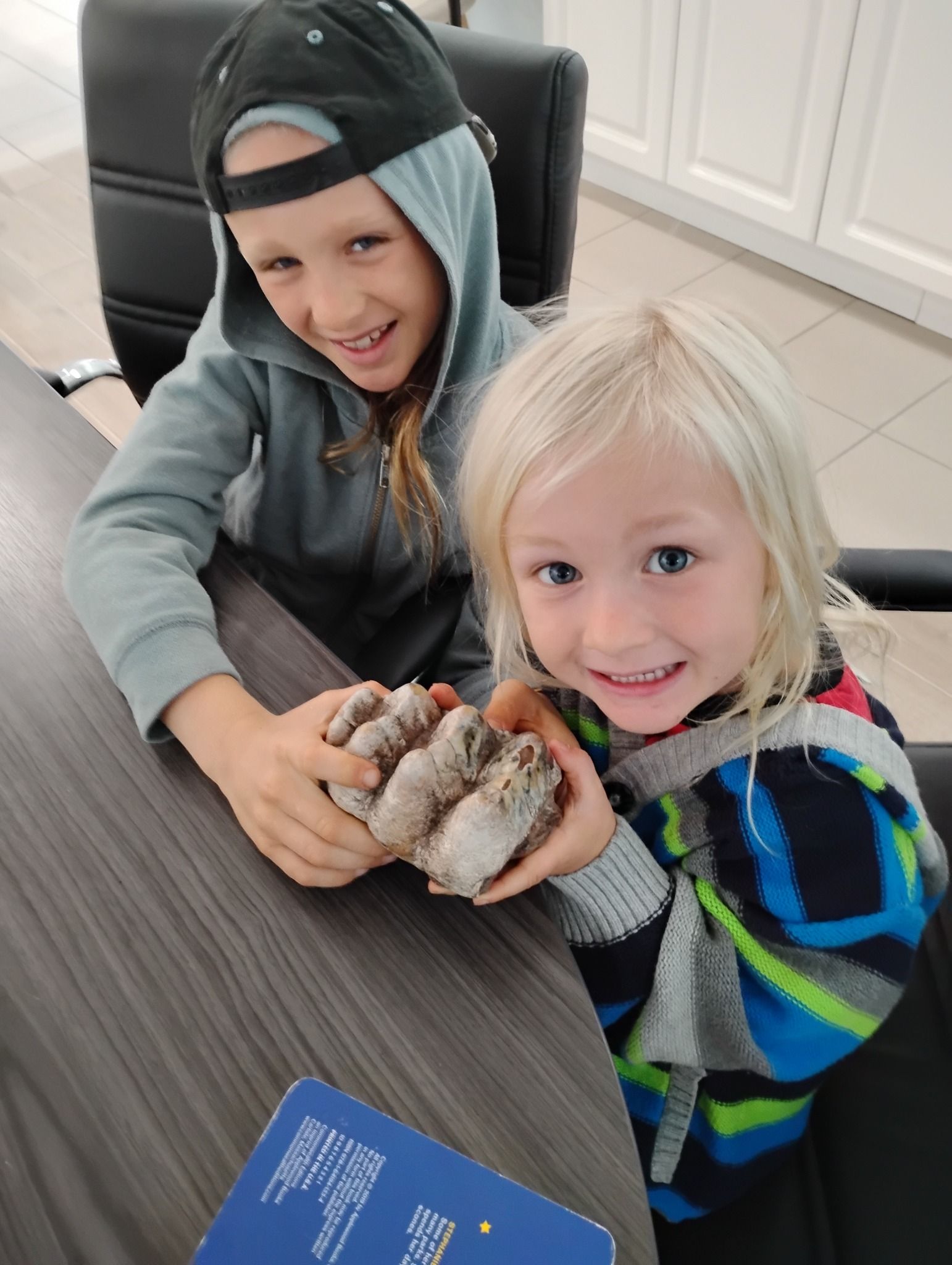 Two children smiling and holding a small hamster at a table indoors