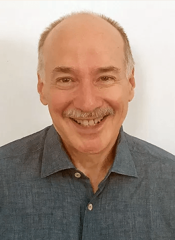 A bald man with a mustache is smiling for the camera while wearing a blue shirt.