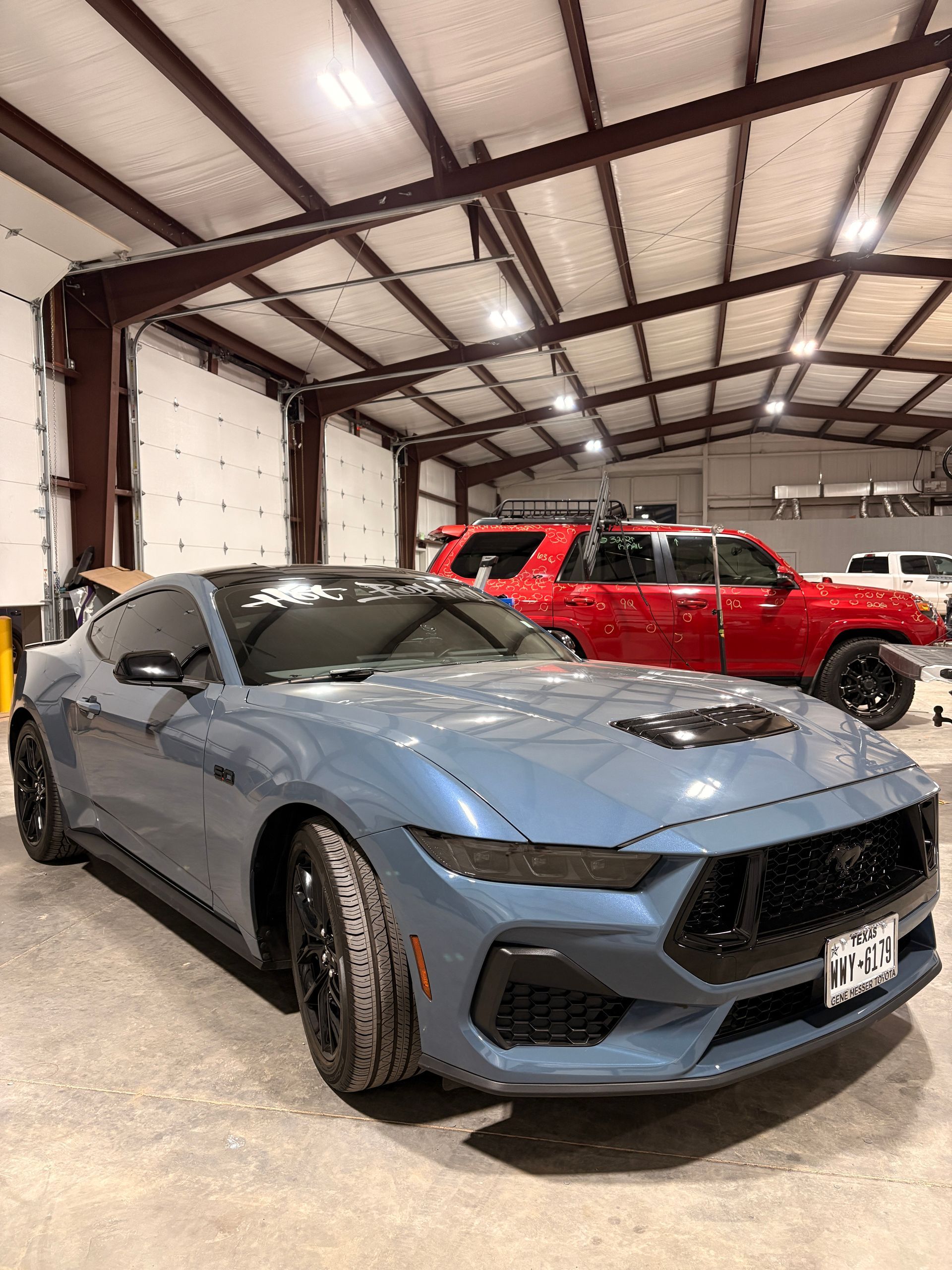 Blue sports car in a warehouse garage with a red SUV parked behind it.