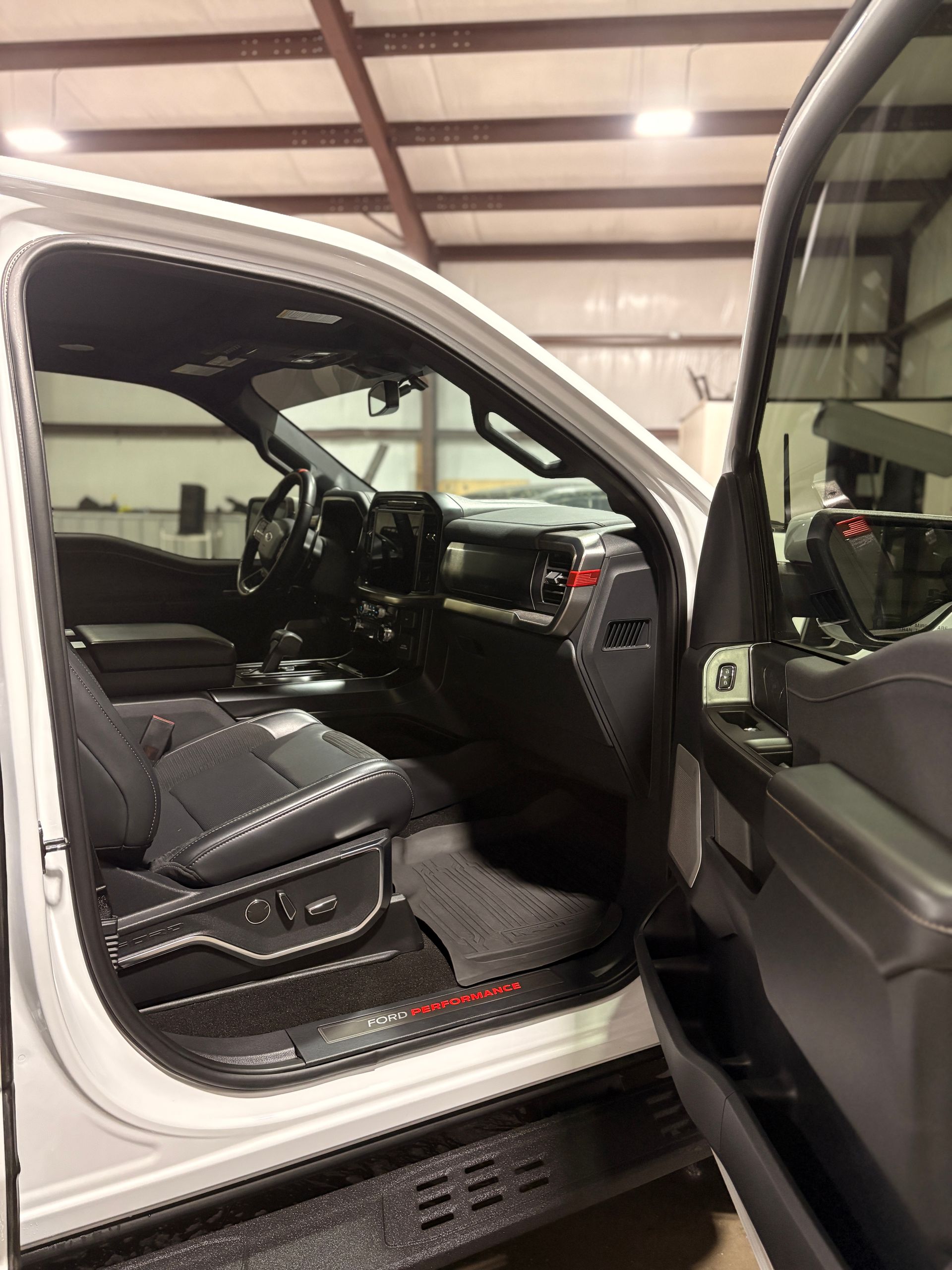 Open car door showing black leather driver seat and dashboard inside a white vehicle in a garage
