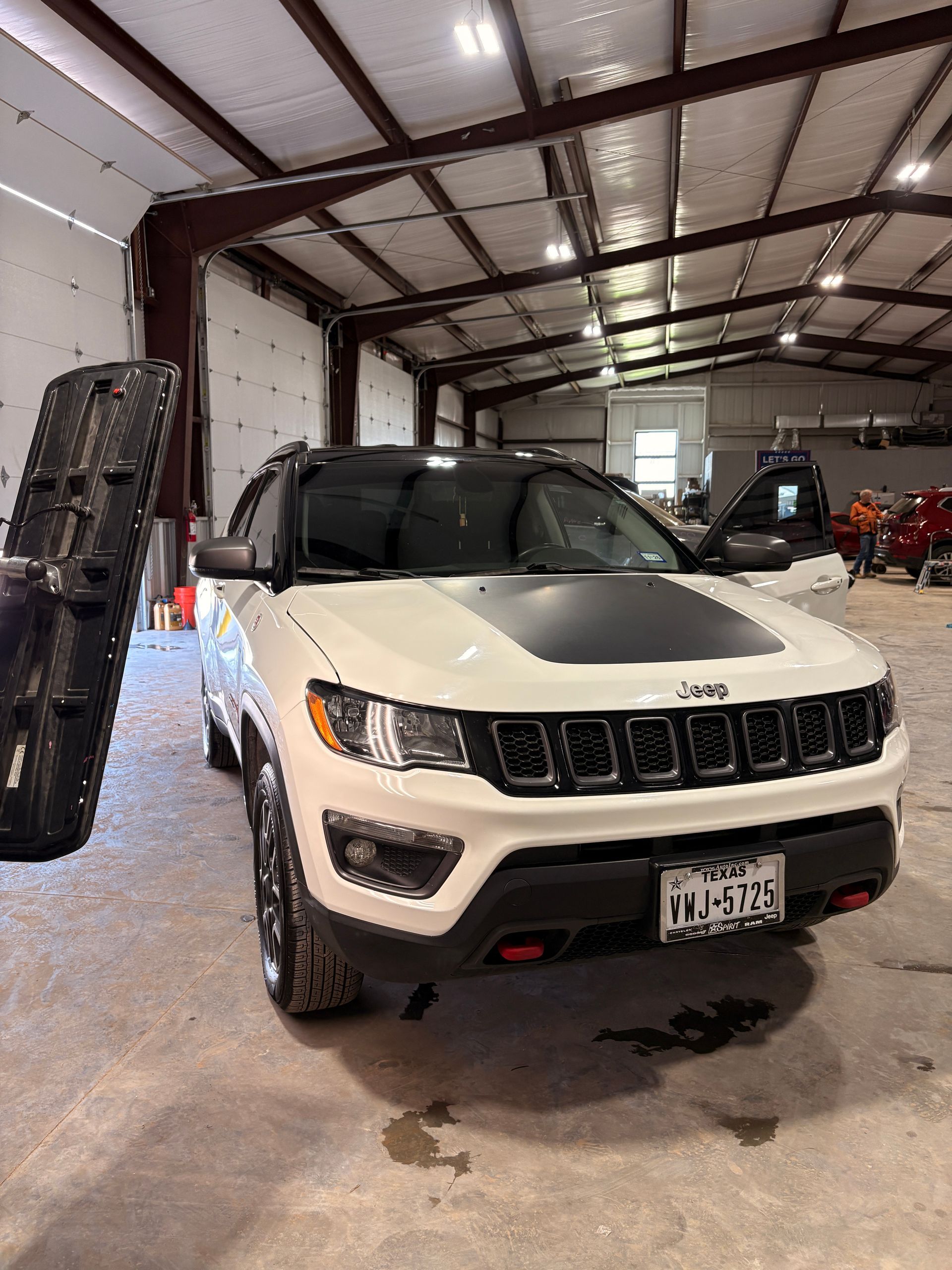 White Jeep SUV in a garage, front view with black hood decal and red tow hooks.