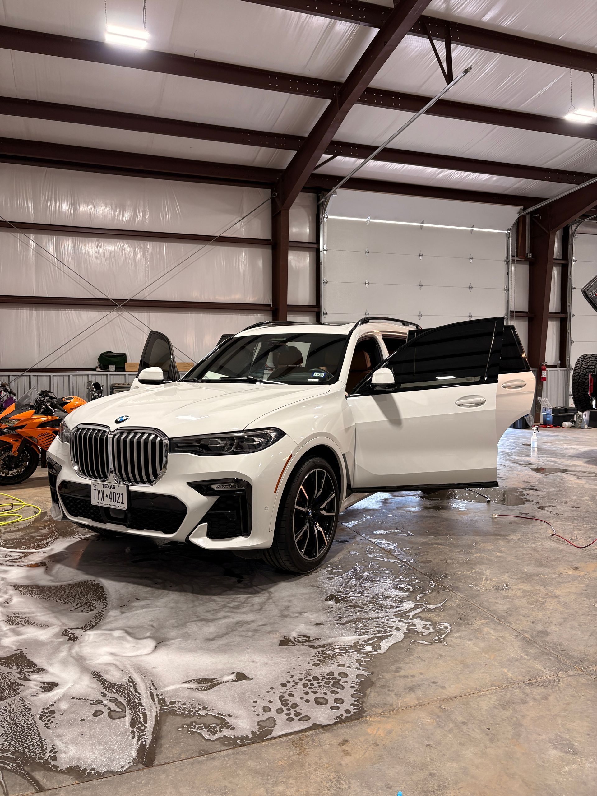White BMW SUV in a garage, driver door open, black cargo on rear seat, wet concrete floor.