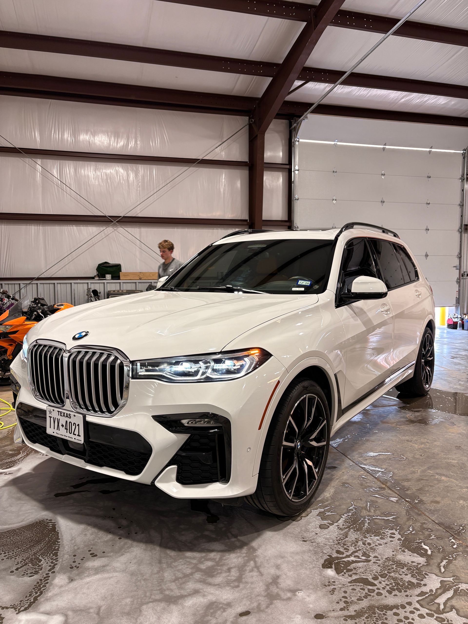 White BMW SUV in a warehouse, viewed from the front-left with wet floor reflections.