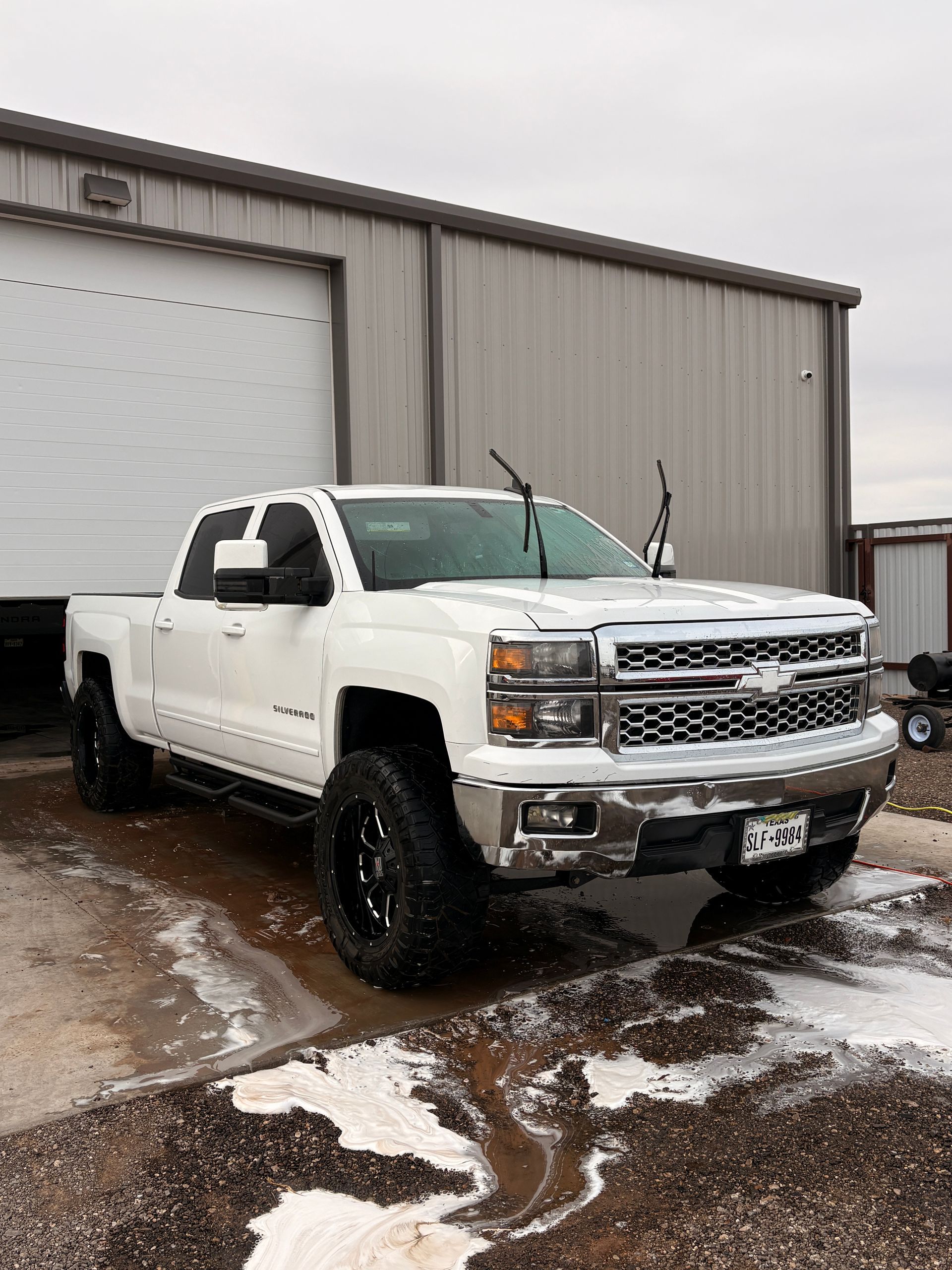 White lifted pickup truck parked outside a metal garage on a snowy lot