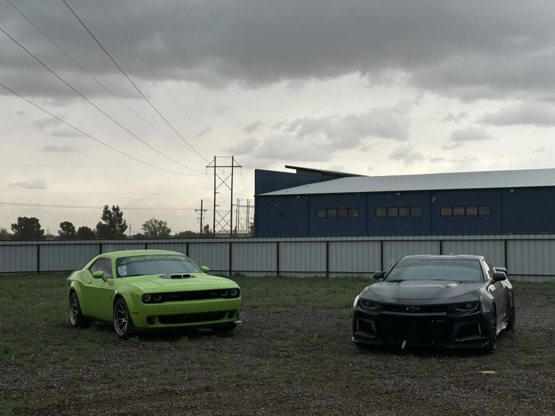 Two sports cars, lime green and black, parked on gravel under a cloudy sky near an industrial building