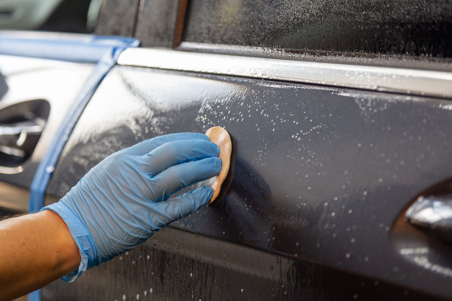 Gloved hand wiping a black car door with a sponge, cleaning soap suds on the surface