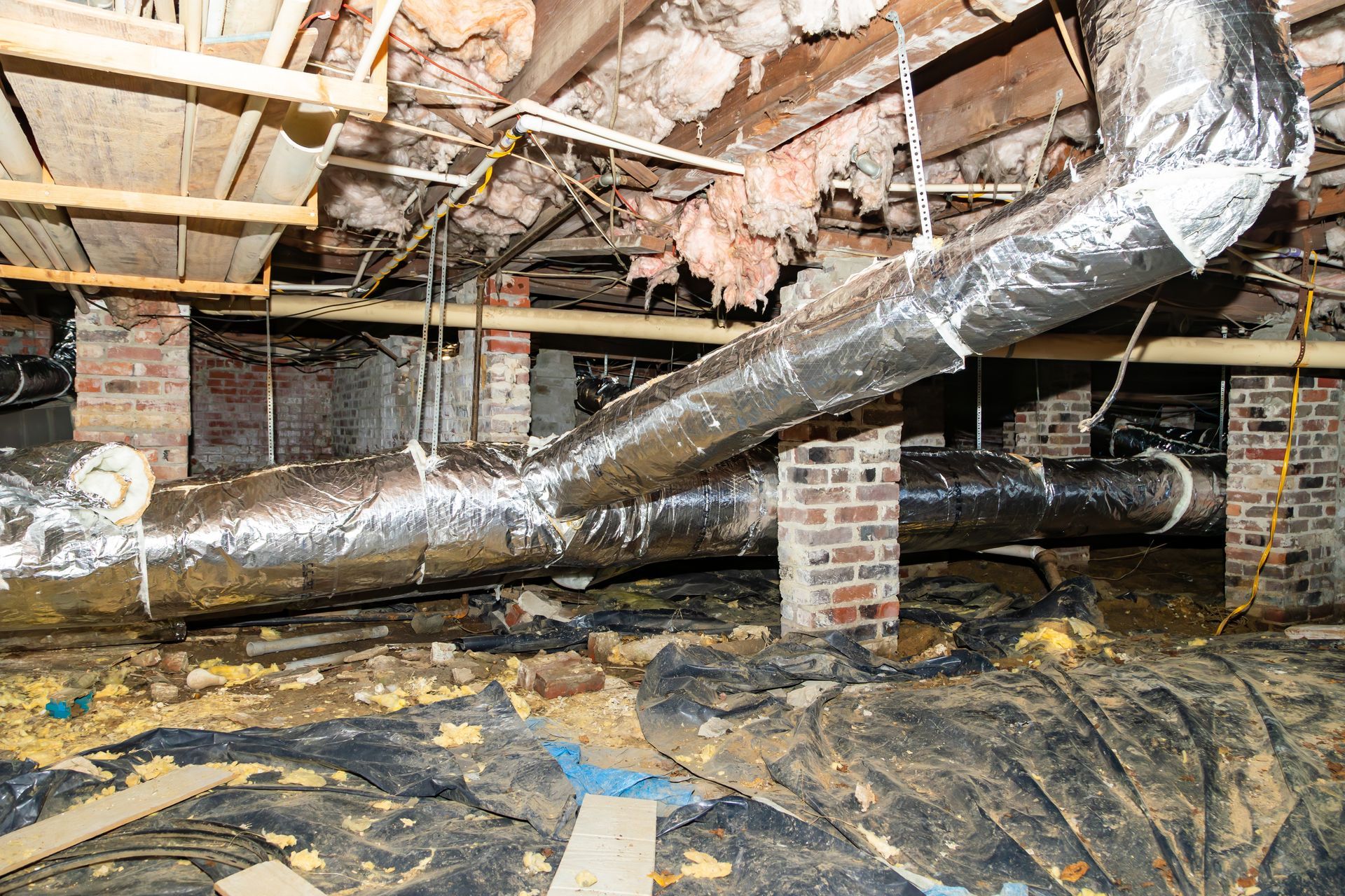 Crawlspace with HVAC ductwork, brick supports, insulation, and debris on the ground.