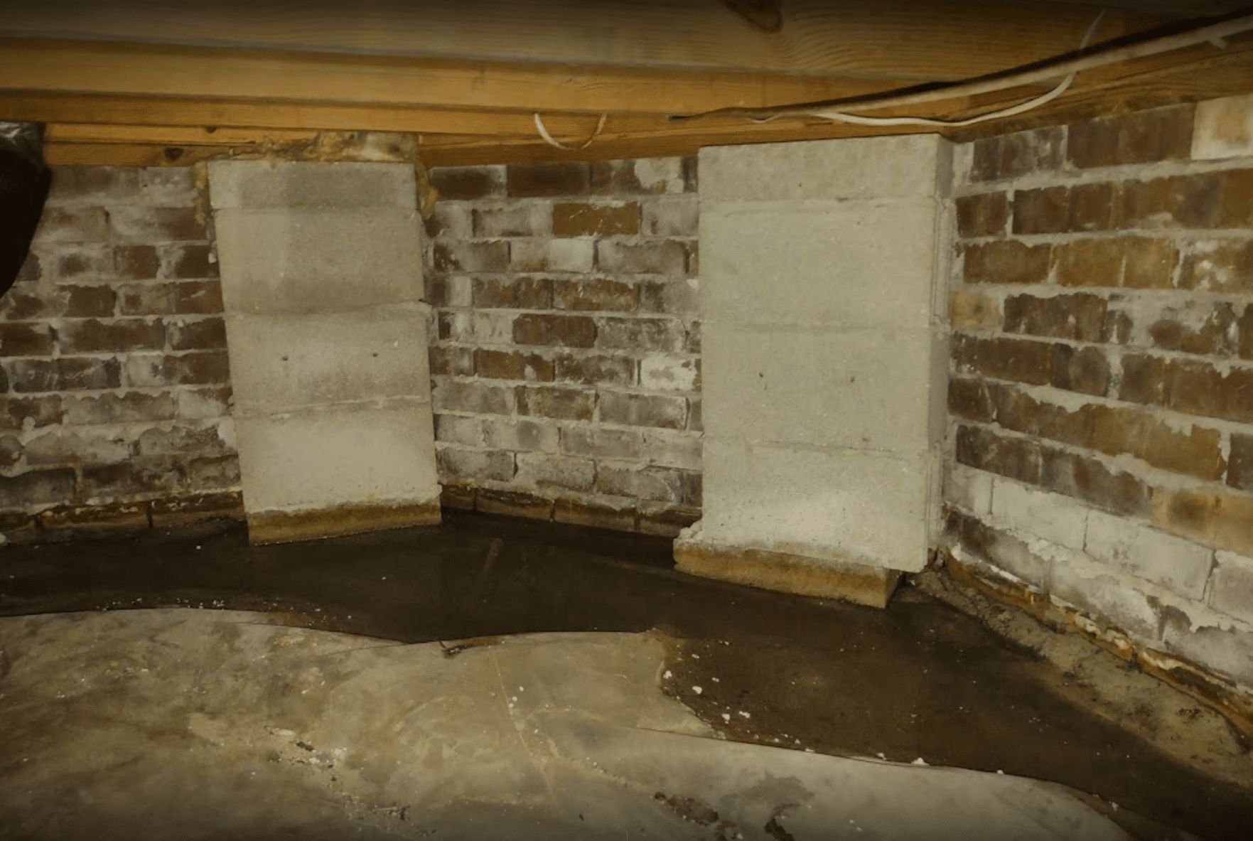 Dark brick basement corner with two white support pillars and a damp concrete floor