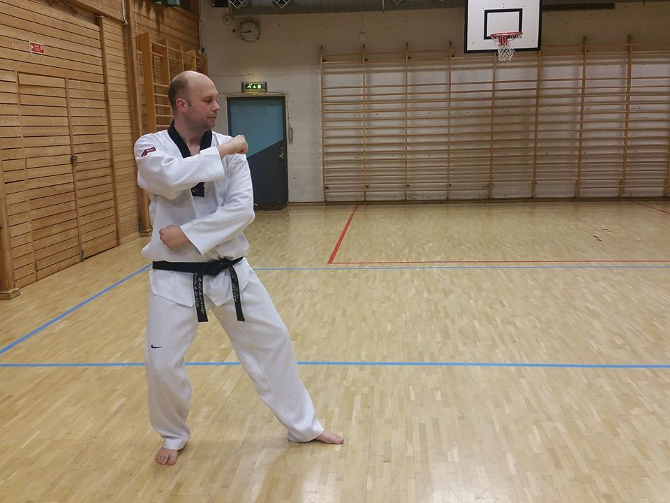 Man in white martial arts uniform performing a block, indoor gym with wooden floor.