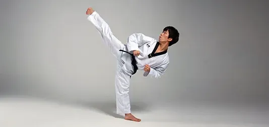 A person in a white martial arts uniform performing a high kick, in a studio with a gray background.