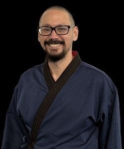 Man in a dark blue martial arts uniform with glasses, smiling in front of a black backdrop.
