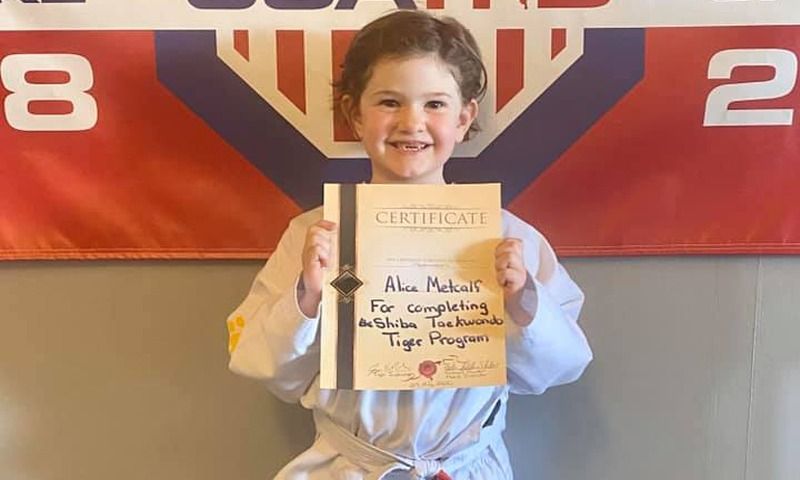 Young child in karate uniform holding a certificate, smiling, against a banner.