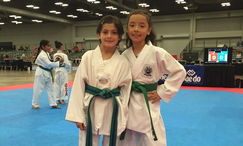 Two girls in green belts and Taekwondo uniforms pose at a tournament, other competitors in background.