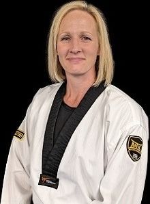 Woman in martial arts uniform with black belt, smiling, against a black background.
