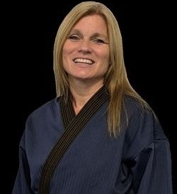 Woman in blue martial arts uniform with black belt, smiling against a black background.
