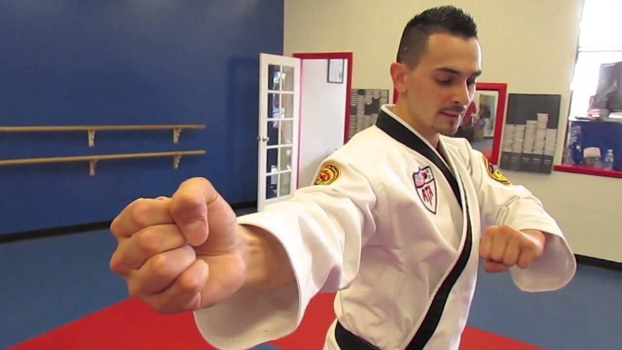 Martial artist in white uniform demonstrates a punch, indoors with blue walls and a red and white door.