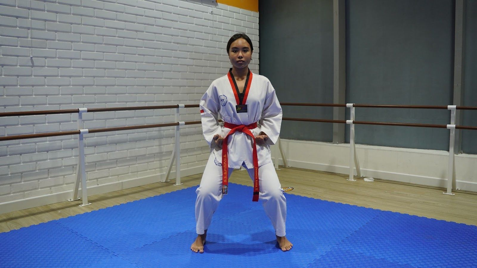 Woman in a red belt and white martial arts uniform, in a ready stance, on blue mats.