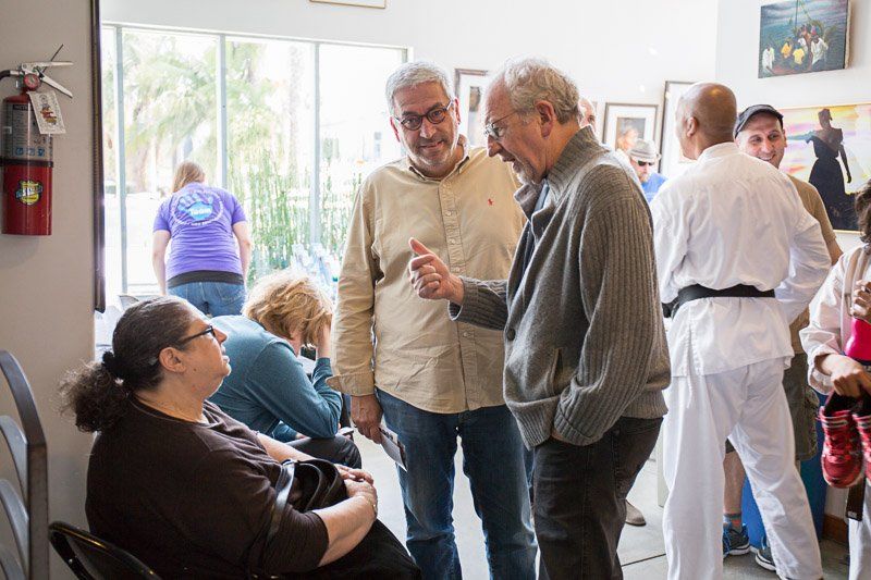 People conversing in a bright room. Two men are speaking, others listen. A woman sits, and a martial artist is in the background.