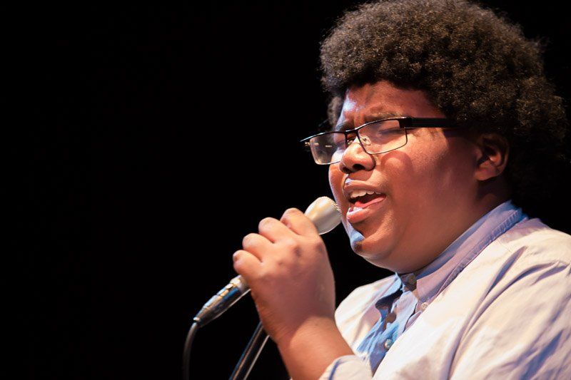 Person with curly hair and glasses singing into a microphone on stage.