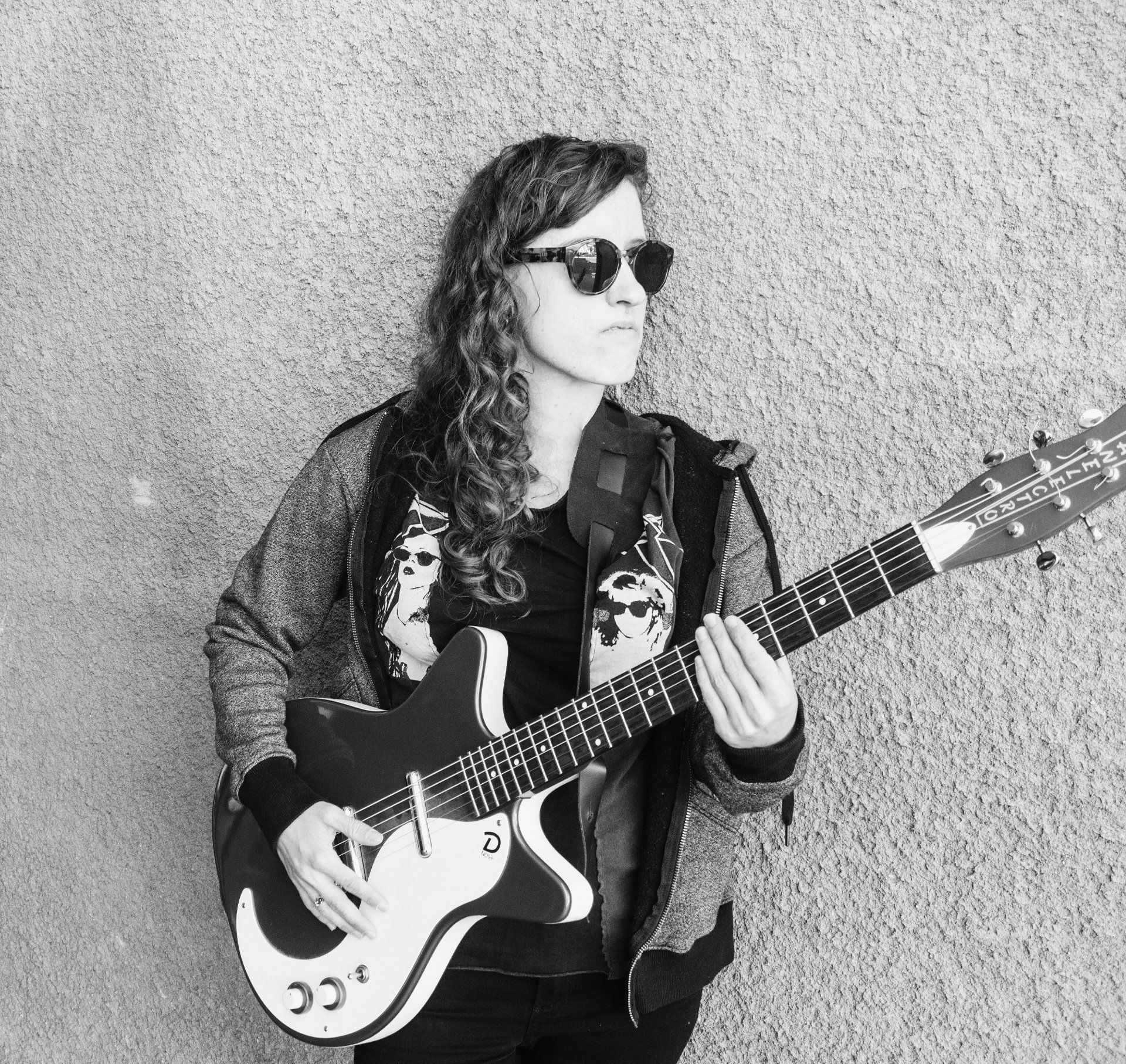 Woman with sunglasses, long hair, holding and playing a white electric guitar against a textured wall.