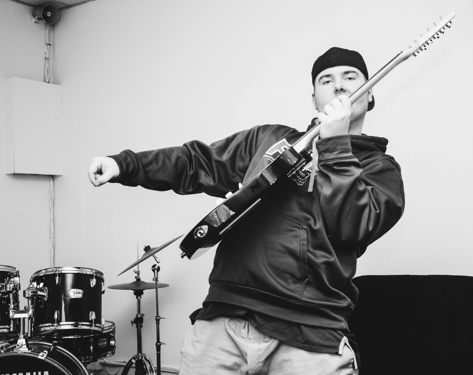 Man in hoodie with guitar, making a fist, near a drum set. Black and white.