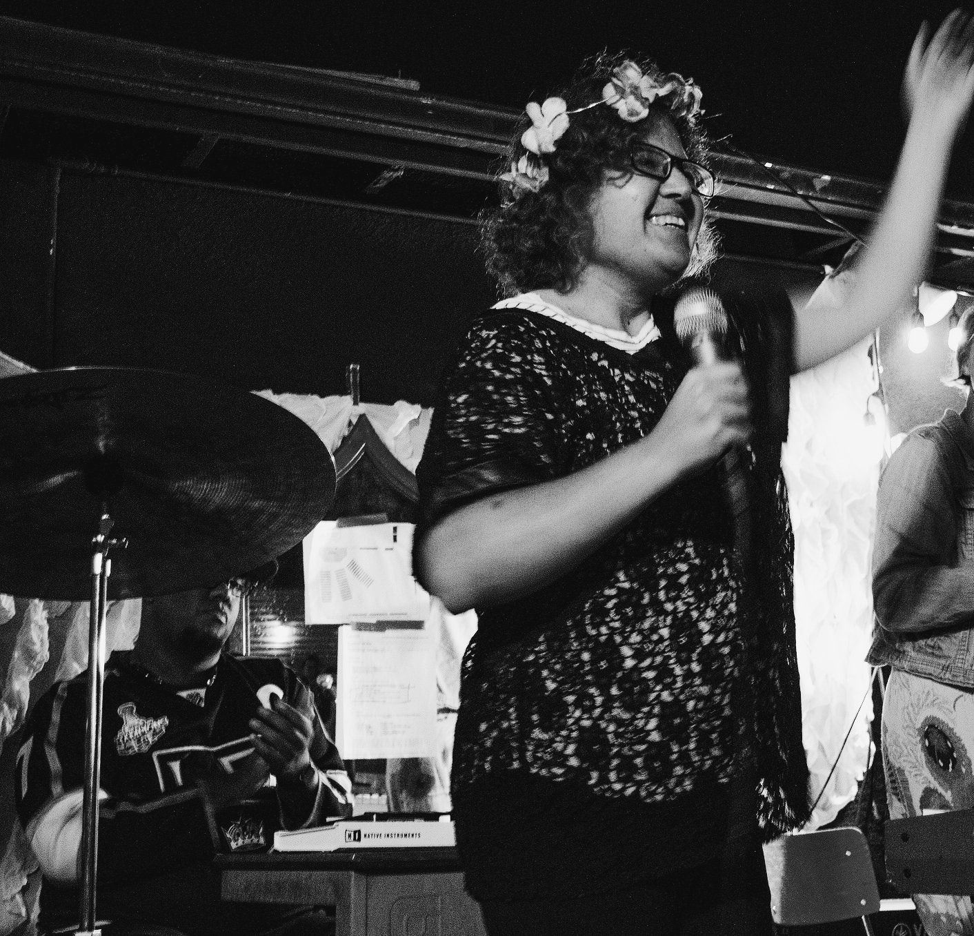 Black and white photo of a person with glasses and a floral crown singing into a microphone, arm raised, on stage. A drummer is in the background.