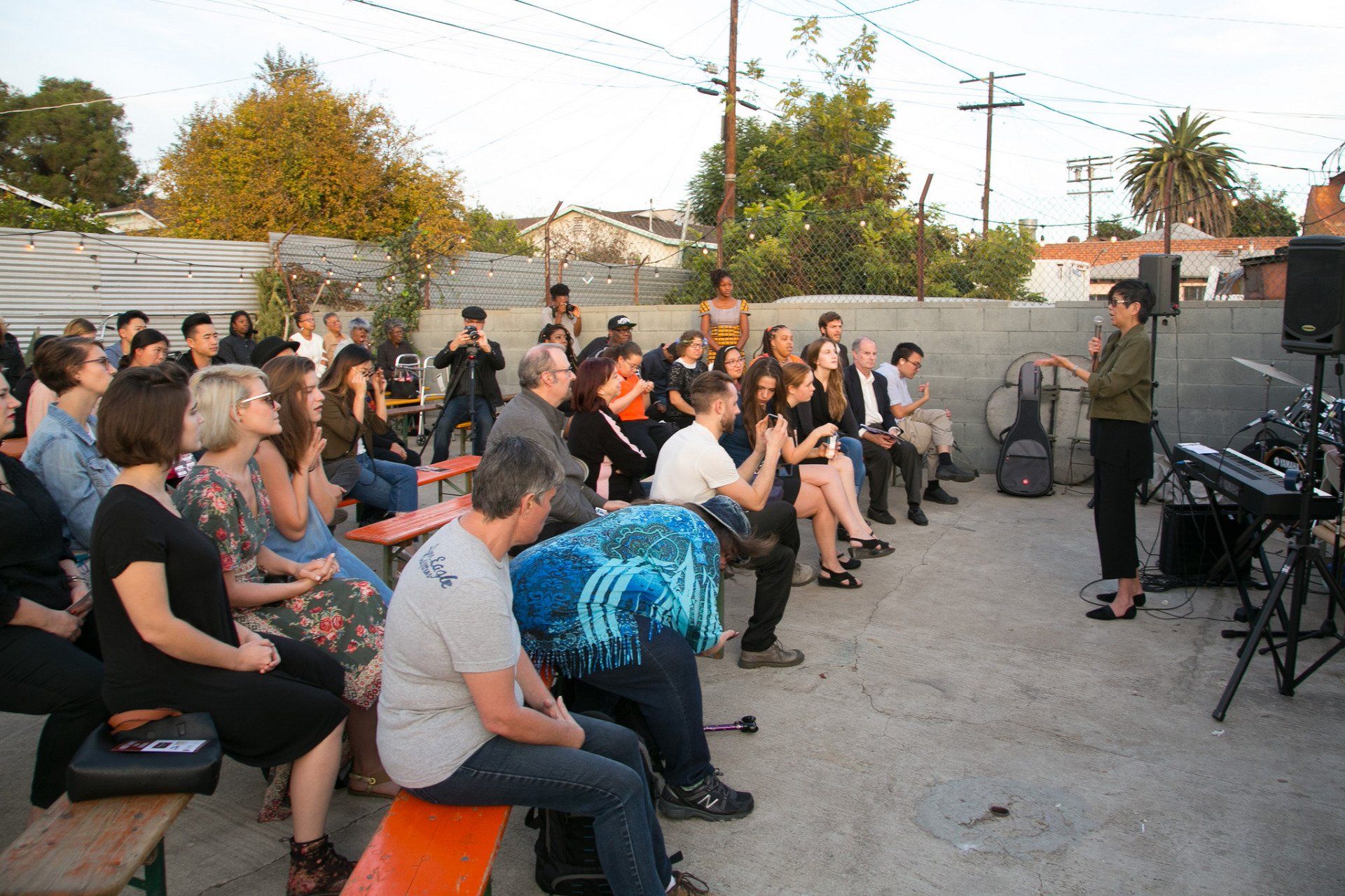 Outdoor event: Woman speaking to a seated audience. People clapping, trees and sky in the background.