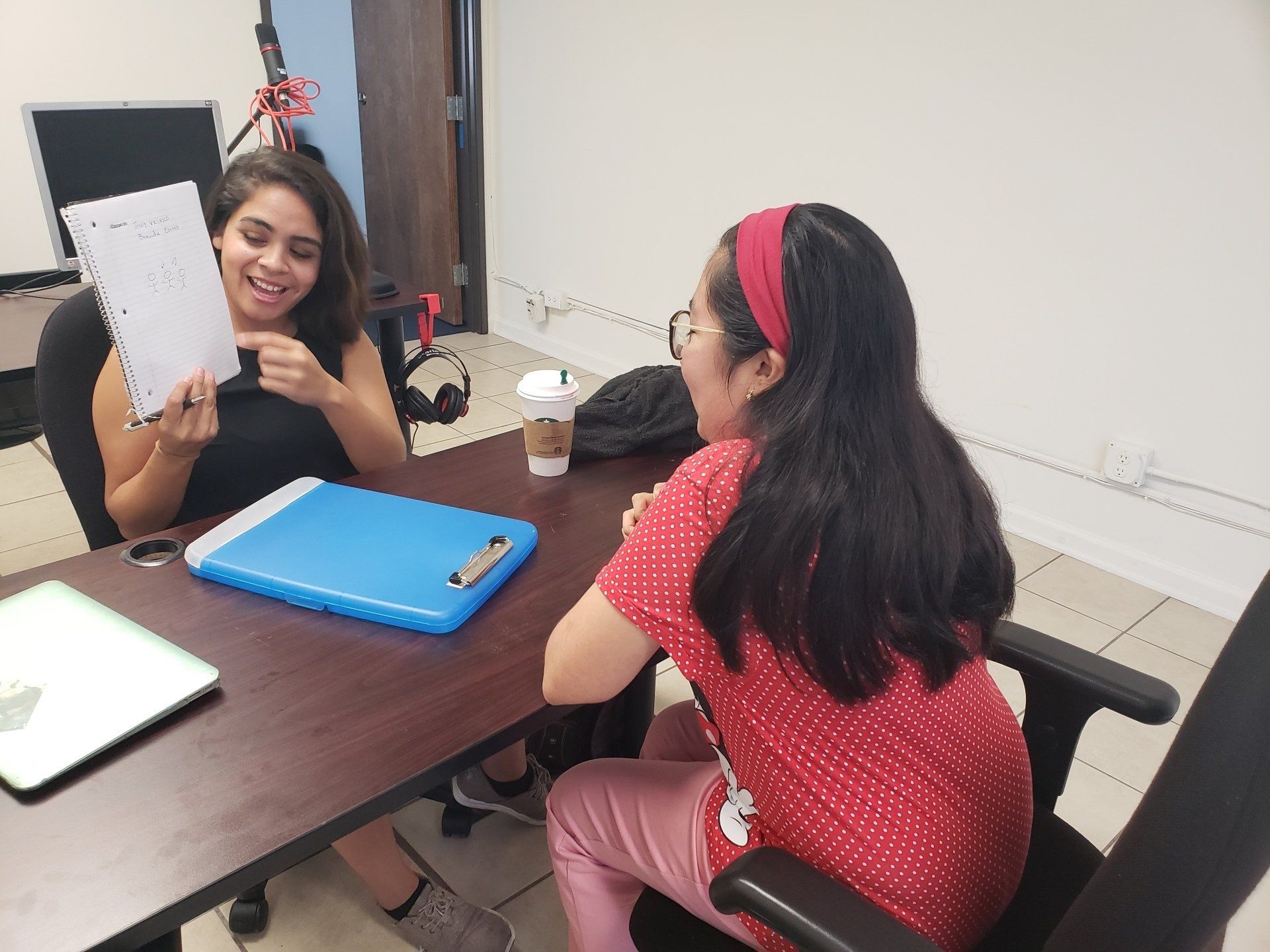 Two women in an office, one showing a paper, both smiling. Desk with laptop, clipboard, and coffee cup.