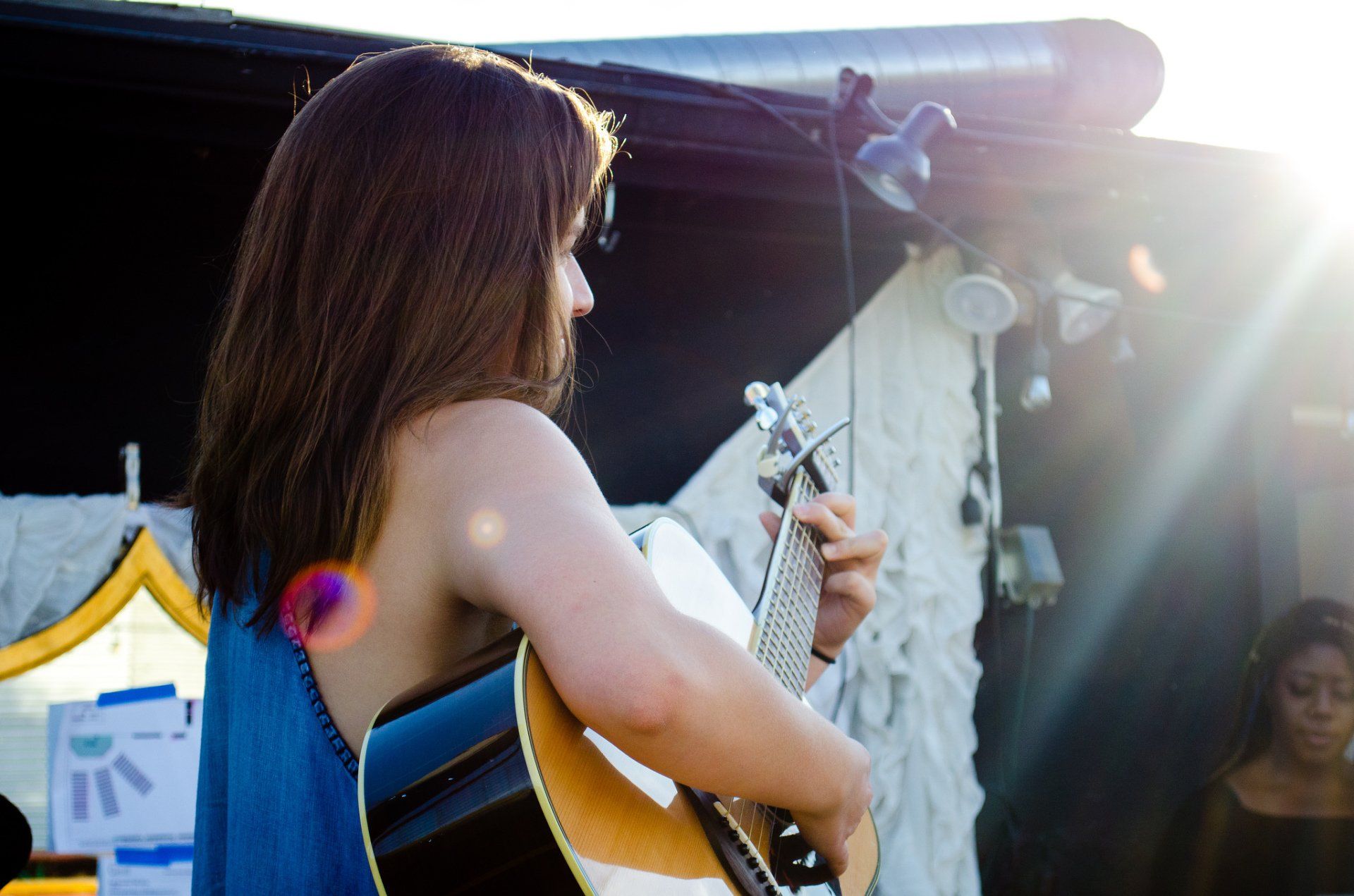 Woman playing acoustic guitar outdoors with sunlight; blue dress, brown hair.