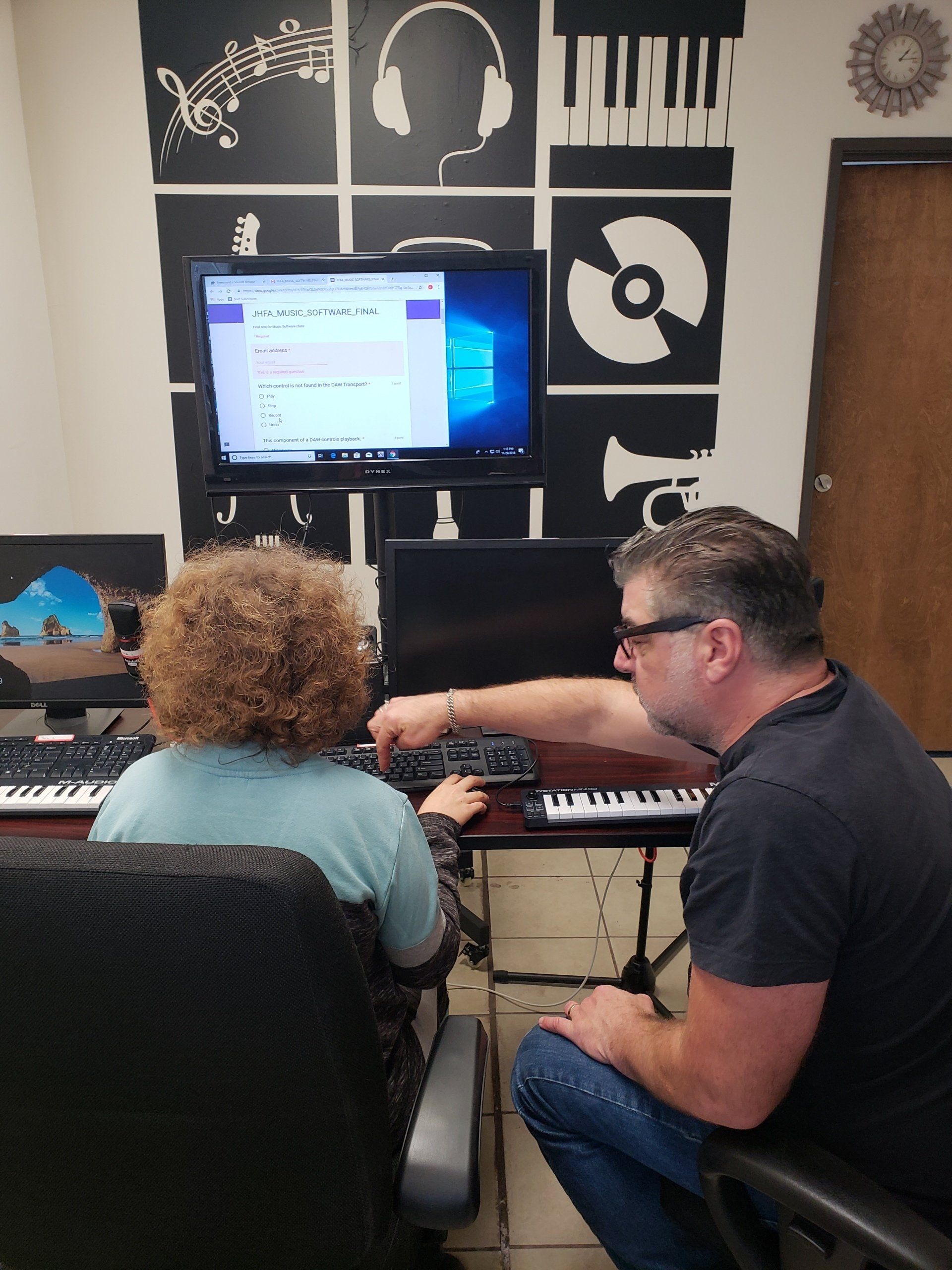 A man coaches a woman at a computer, in a music studio; black and white decor.