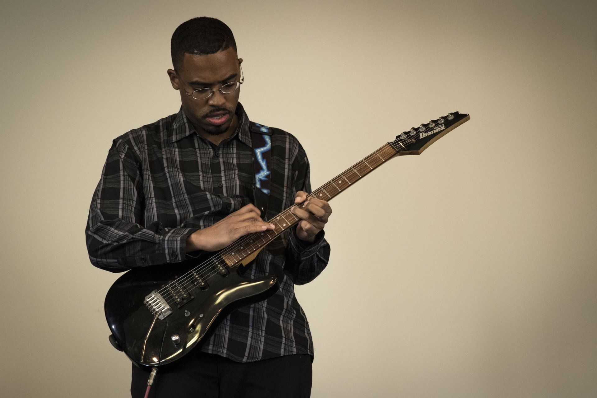 A Black man wearing glasses plays an electric guitar against a beige backdrop.