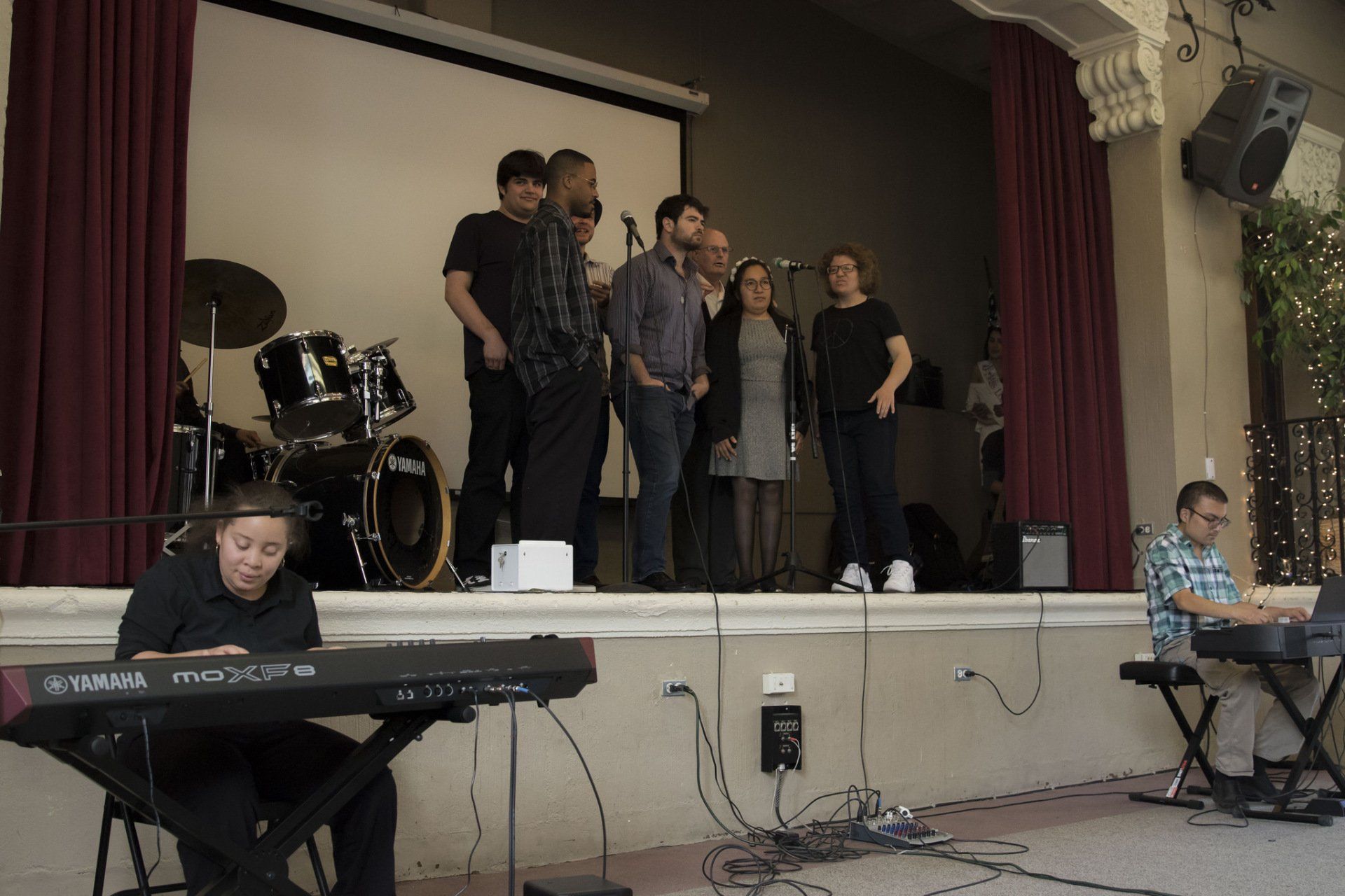 A band performs on stage; two keyboardists in front, a drummer in the back, and singers with a microphone.