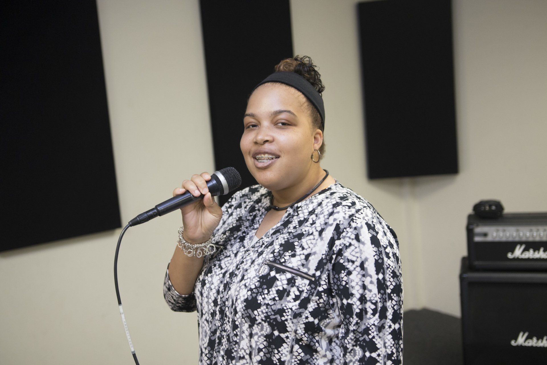 Woman with microphone, singing in a studio. Wearing patterned top, headband, and jewelry. Two black sound panels are in the background.