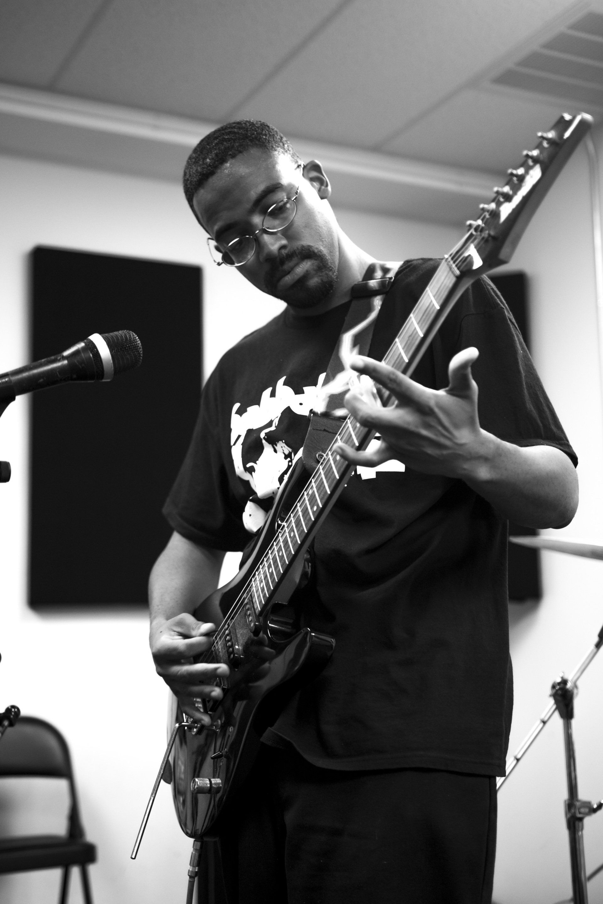 Black and white photo of a man playing an electric guitar. He's wearing glasses and a t-shirt, in a music studio setting.