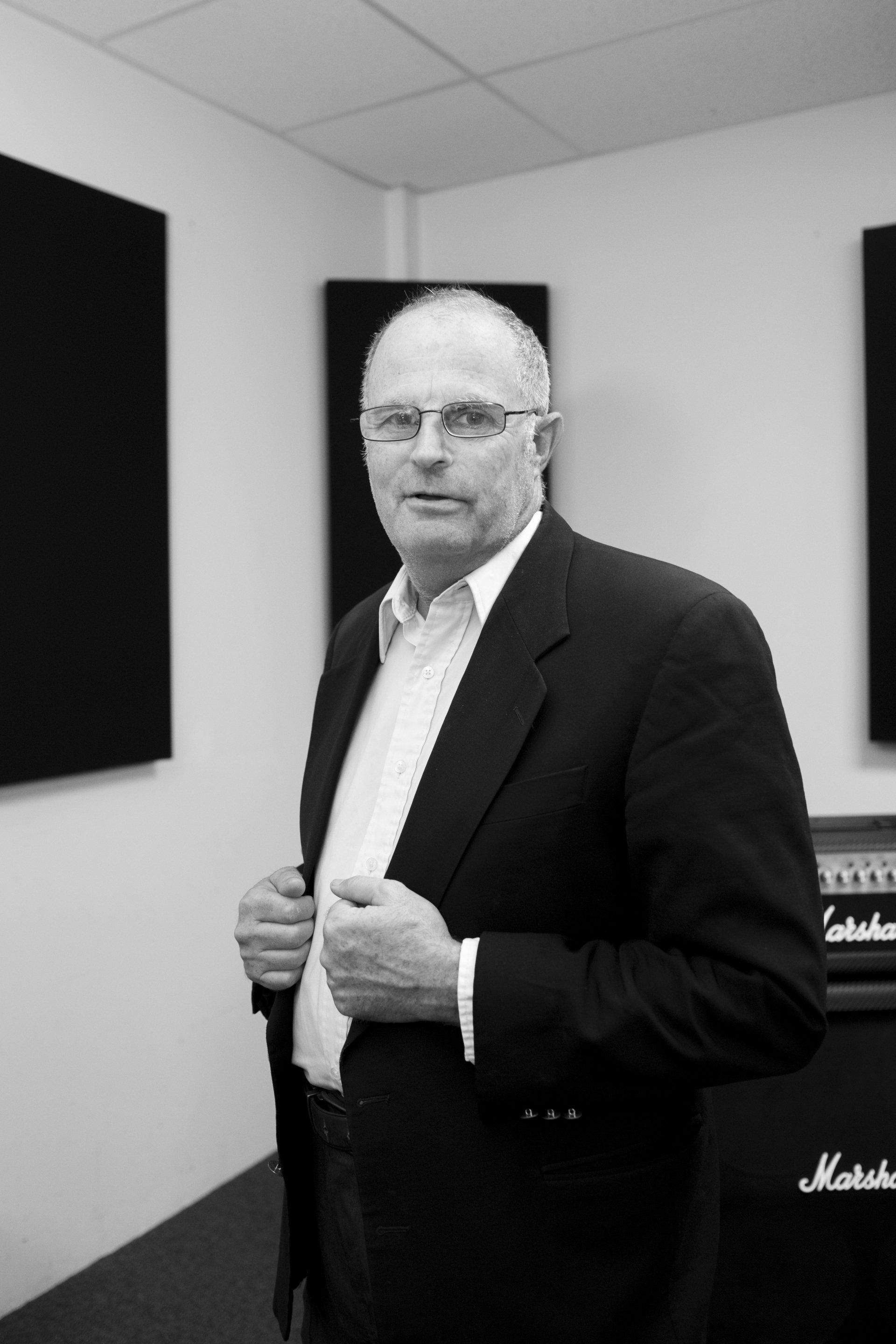 Man in blazer, holding it open. He’s in a room with studio soundproofing and an amp.