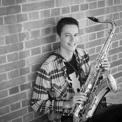 Young man smiles, holding a saxophone, leaning against a brick wall.