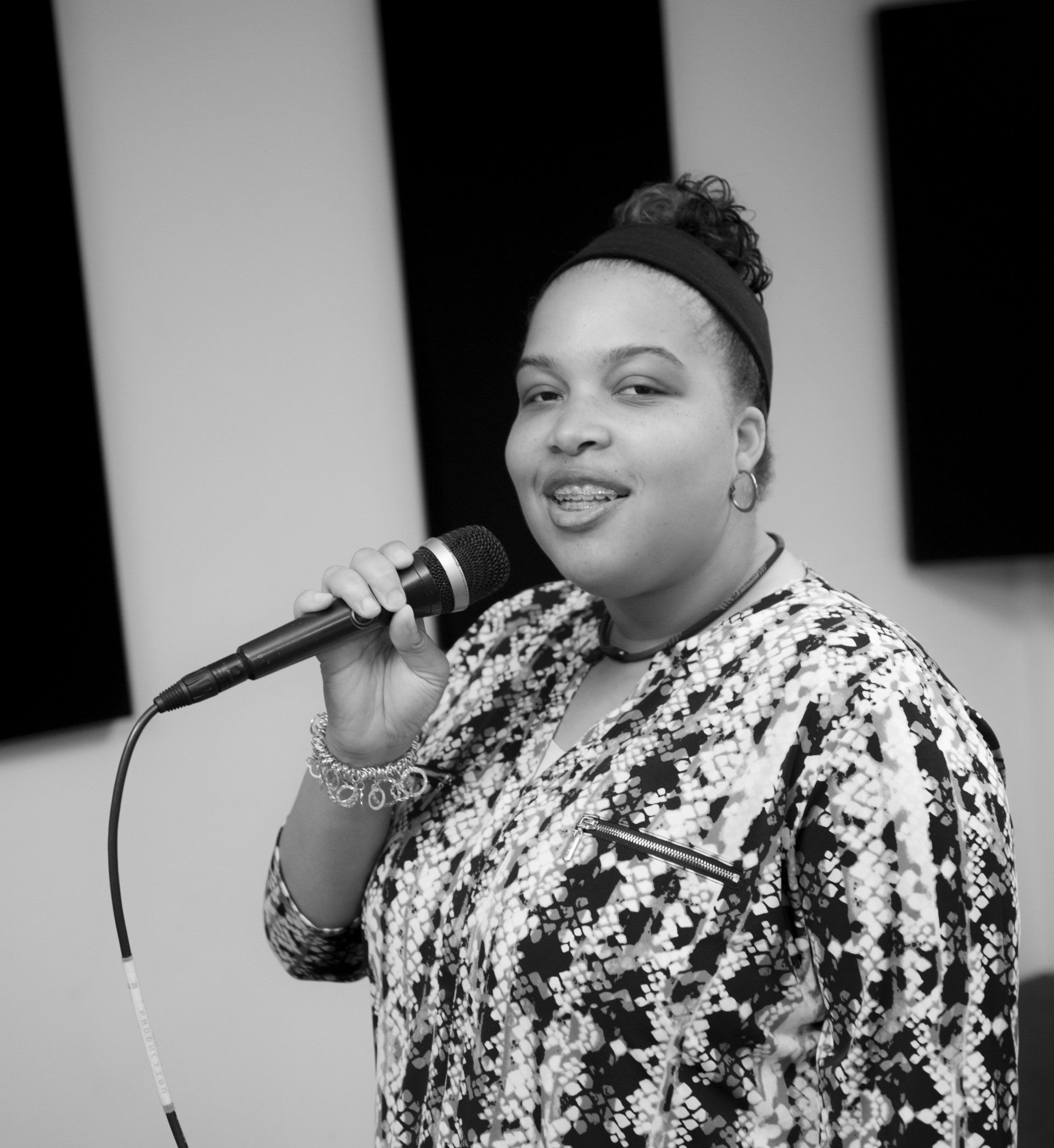 Woman singing into a microphone, wearing a patterned shirt. Indoors, black and white.