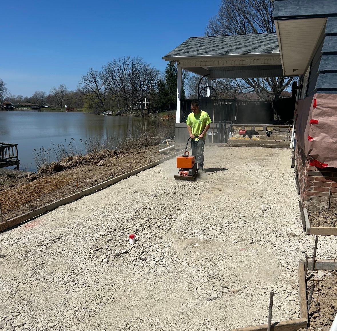 A Lavoie Concrete worker uses a plate compactor on a gravel area next to a house and a lake.