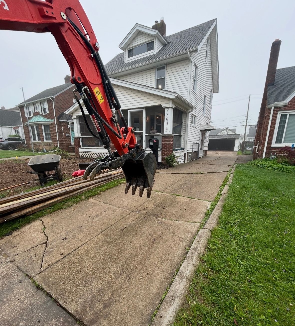 A red excavator boom and bucket hover over a residential concrete driveway in front of a white two-story house.