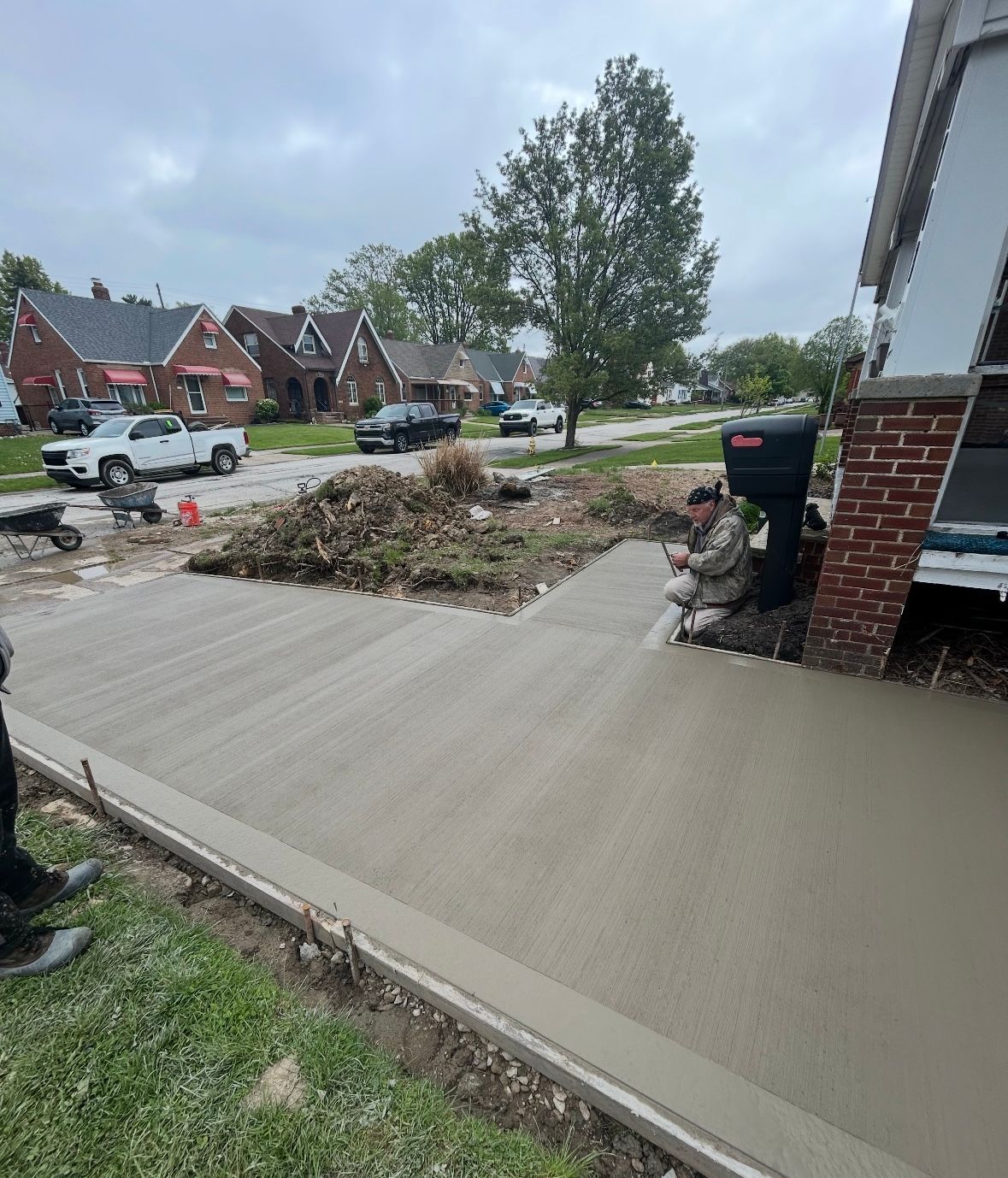 A person finishes smoothing a freshly poured concrete driveway in front of a brick house on a residential street.