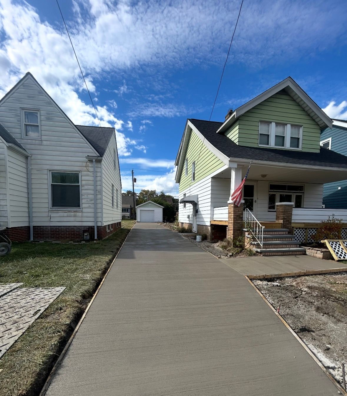 A newly poured concrete driveway leads between two houses toward a detached garage under a blue, cloudy sky.