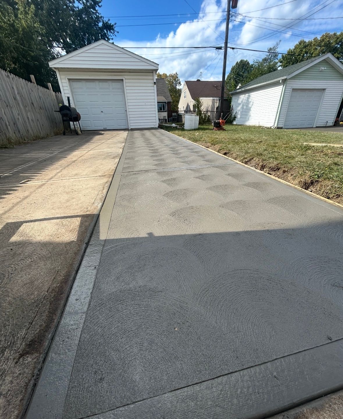 A newly paved gray concrete driveway section running alongside an older, weathered concrete driveway toward two garages.