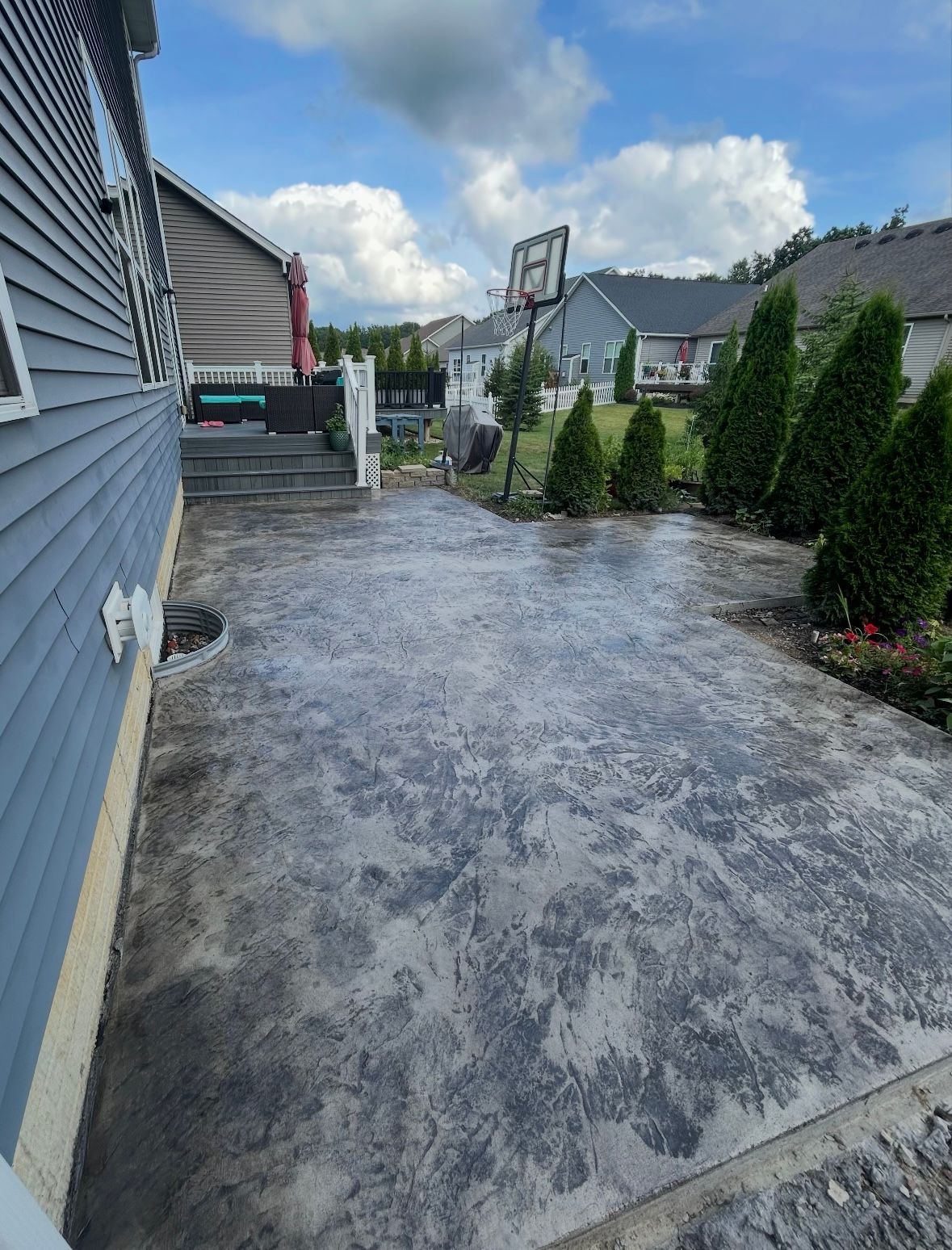 A stamped, textured concrete patio runs along the side of a blue-sided house toward a backyard with a basketball hoop.