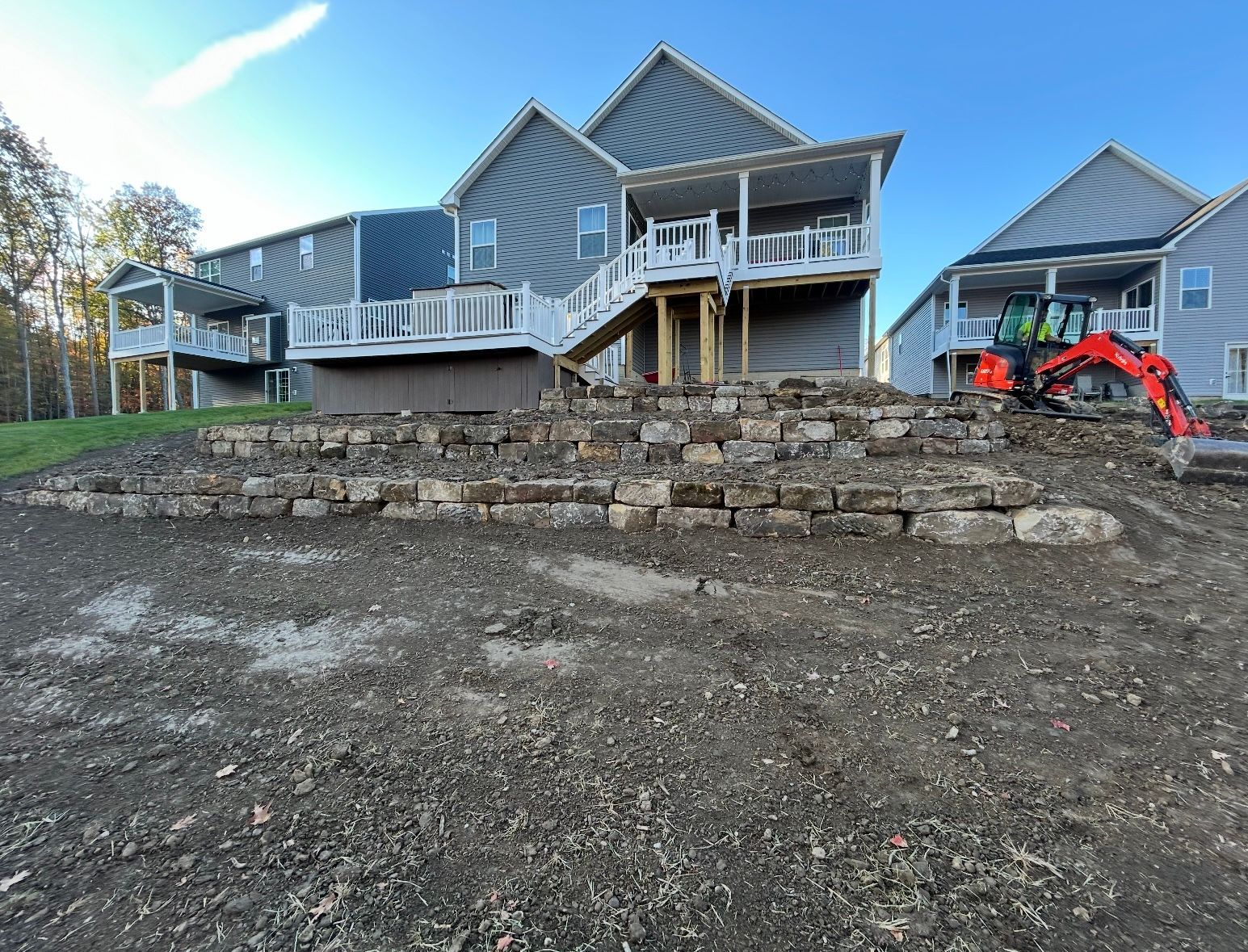 An orange excavator works on a tiered stone retaining wall in the backyard of a multi-story gray house under a blue sky.