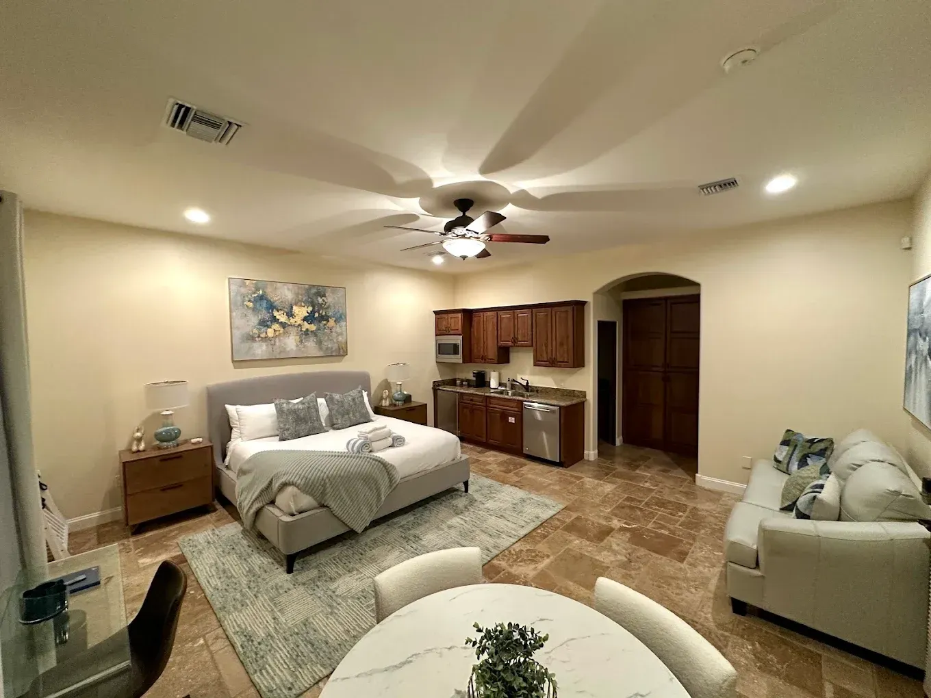 Cozy studio apartment with a bed, kitchenette, sofa, and dining table, beige walls, and brown tile floor.