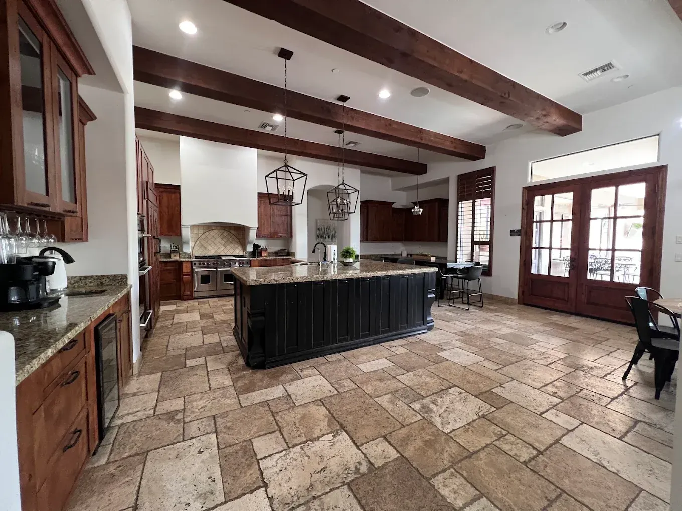 Spacious kitchen with dark island, wooden cabinets, and stone tile flooring. Dark beams across ceiling.