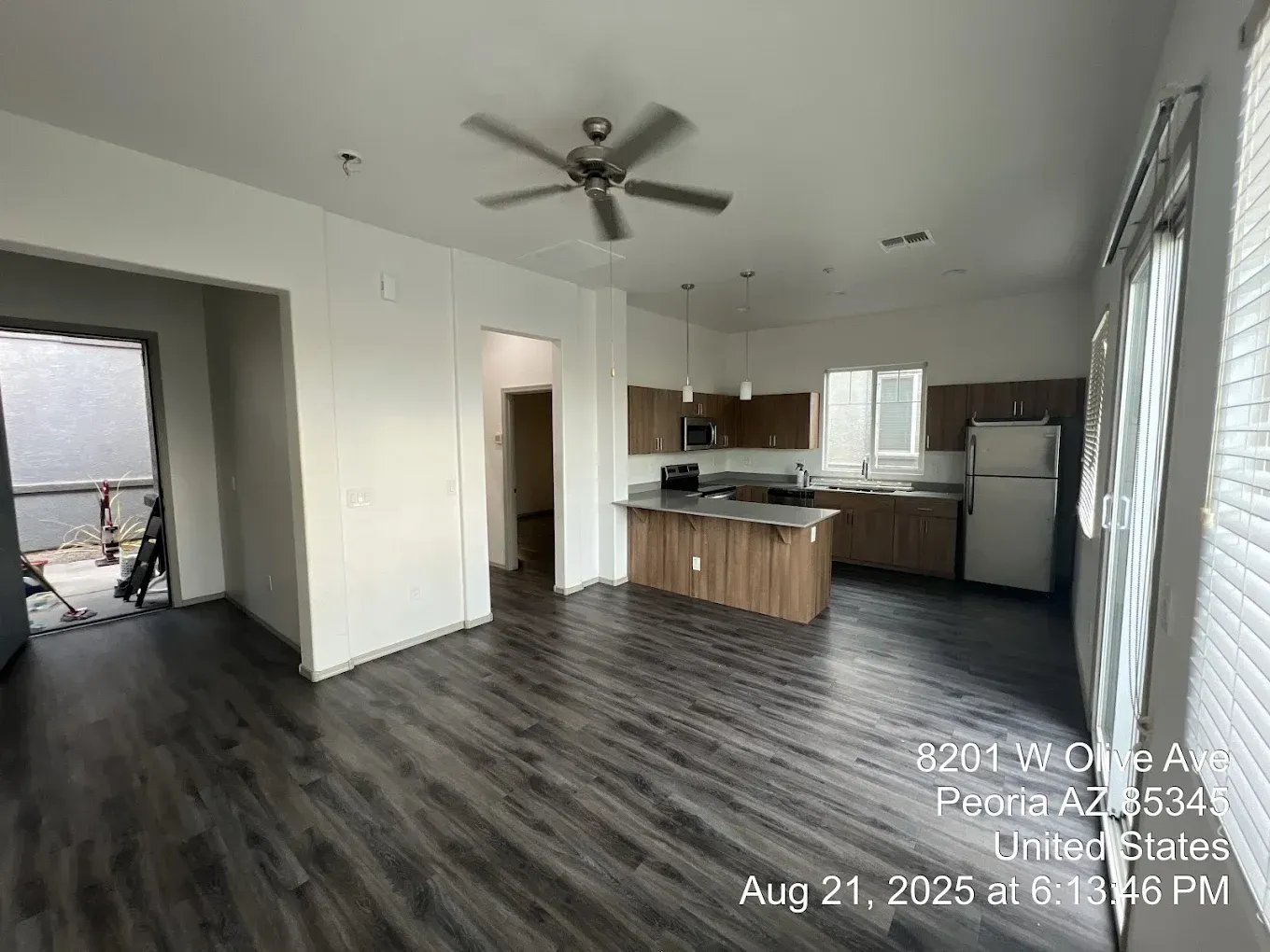 Open-concept living space with kitchen. Dark wood flooring, light walls. Kitchen island, appliances, ceiling fan.