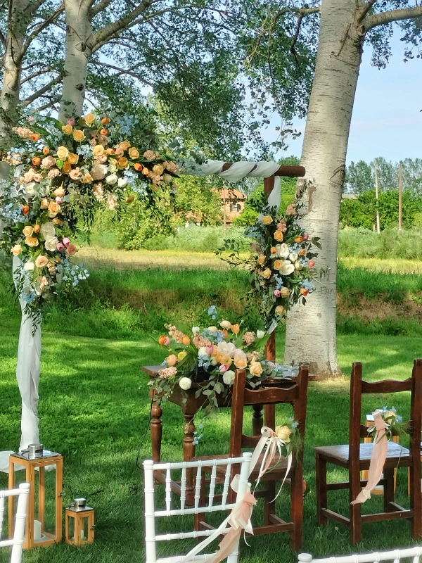 Allestimento per cerimonia nuziale con arco floreale, sedie in legno e tavolo all'aperto in un giardino.