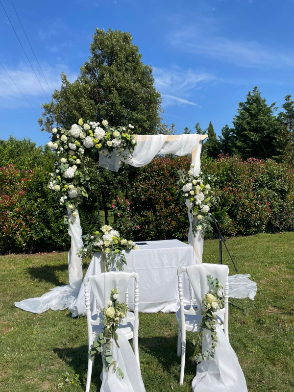 Allestimento per cerimonia nuziale con arco floreale bianco e tessuto drappeggiato in un ambiente esterno soleggiato.